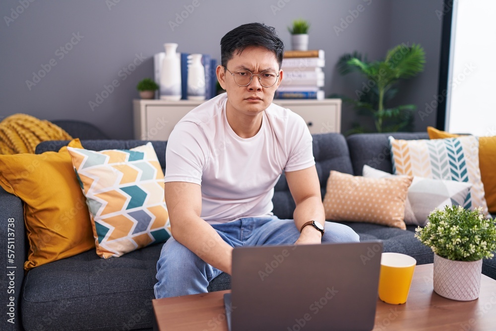 Young asian man using laptop at home sitting on the sofa skeptic and nervous, frowning upset because of problem. negative person. Young asian man using laptop at home sitting on the sofa skeptic and nervous, frowning upset because of problem. negative person.