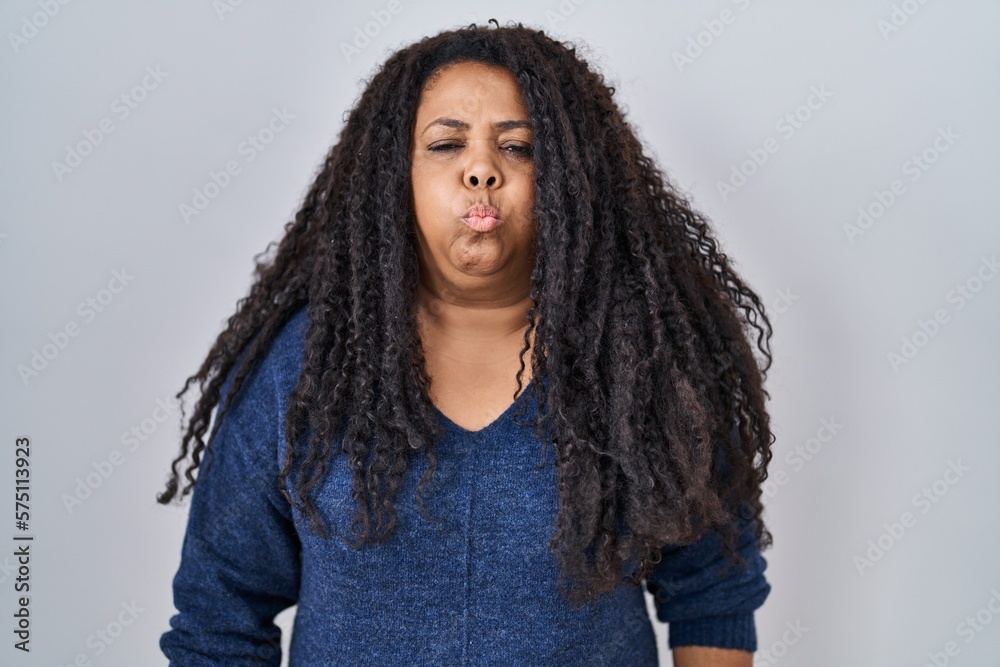 Plus size hispanic woman standing over white background puffing cheeks with funny face. mouth inflated with air, crazy expression. Plus size hispanic woman standing over white background puffing cheeks with funny face. mouth inflated with air, crazy expression.