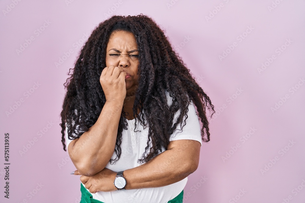 Plus size hispanic woman standing over pink background looking stressed and nervous with hands on mouth biting nails. anxiety problem. Plus size hispanic woman standing over pink background looking stressed and nervous with hands on mouth biting nails. anxiety problem.