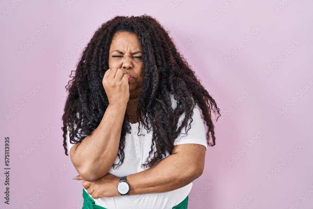 Plus size hispanic woman standing over pink background looking stressed and nervous with hands on mouth biting nails. anxiety problem.