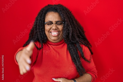 Plus size hispanic woman standing over red background smiling friendly offering handshake as greeting and welcoming. successful business. © Krakenimages.com