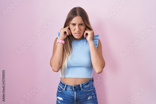 Young blonde woman standing over pink background covering ears with fingers with annoyed expression for the noise of loud music. deaf concept. © Krakenimages.com