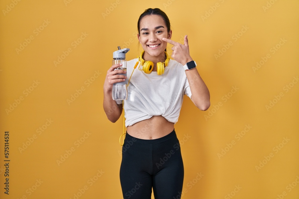 Young south asian woman wearing sportswear drinking water pointing with hand finger to face and nose, smiling cheerful. beauty concept