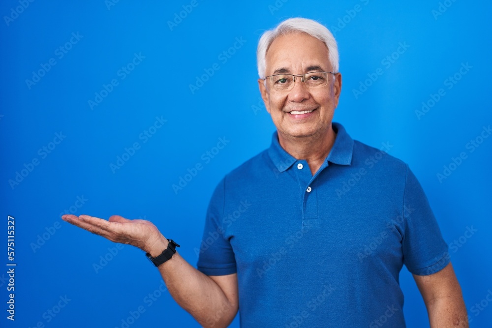 Middle age man with grey hair standing over blue background smiling cheerful presenting and pointing with palm of hand looking at the camera. Middle age man with grey hair standing over blue background smiling cheerful presenting and pointing with palm of hand looking at the camera.