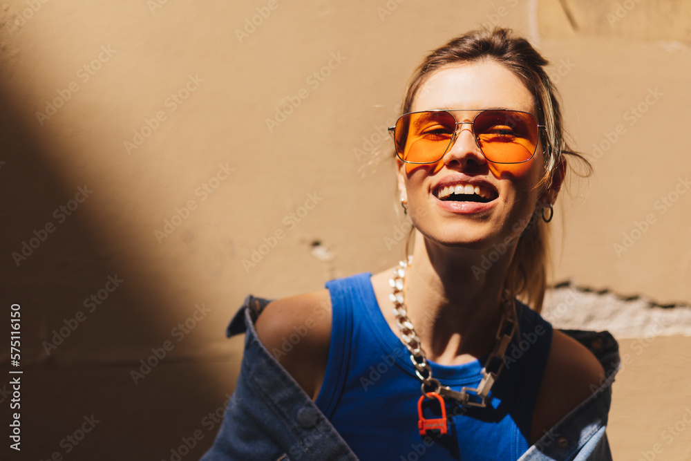Attractive woman with high ponytail standing near beige wall on the street of old city. Girl joy of sun light, wear orange sunglasses look at sun. Shadow and light of sunny summer, spring or fall day. - obrazy, fototapety, plakaty 