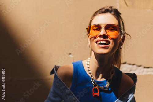Attractive woman with high ponytail standing near beige wall on the street of old city. Girl joy of sun light, wear orange sunglasses look at sun. Shadow and light of sunny summer, spring or fall day.