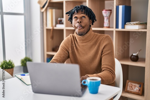 Young african man with dreadlocks working using computer laptop making fish face with lips, crazy and comical gesture. funny expression.