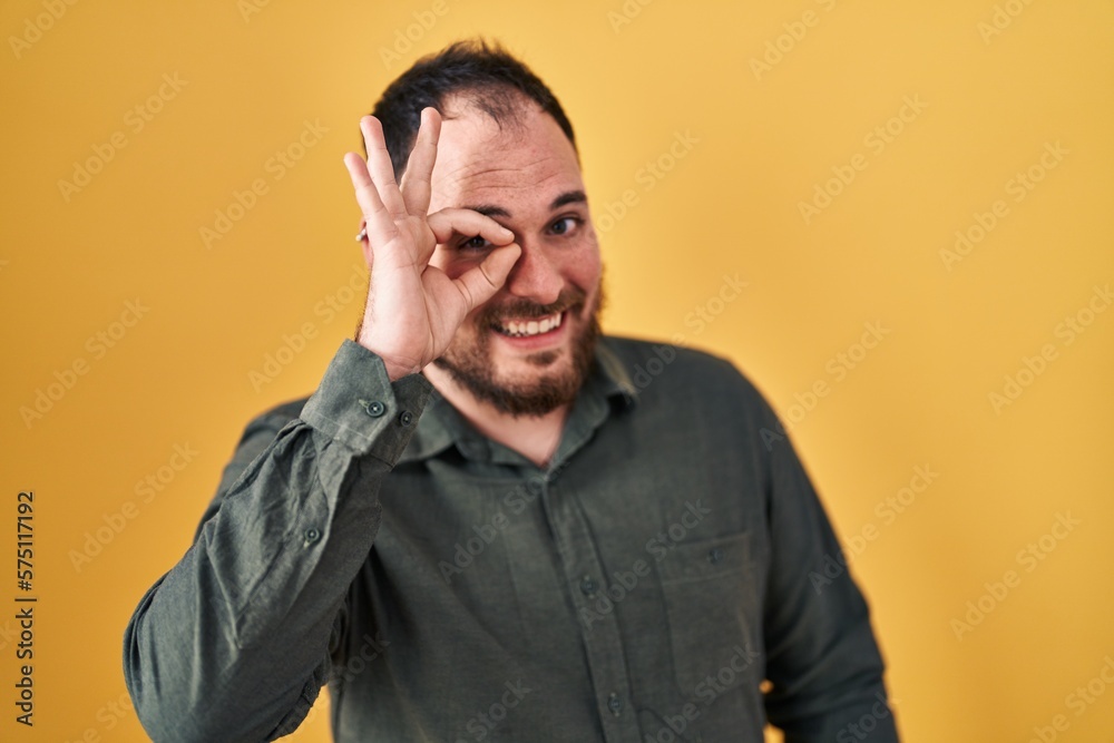 Plus size hispanic man with beard standing over yellow background doing ok gesture with hand smiling, eye looking through fingers with happy face.