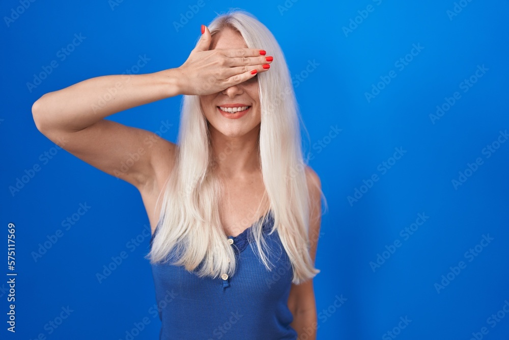 Caucasian woman standing over blue background smiling and laughing with hand on face covering eyes for surprise. blind concept. Caucasian woman standing over blue background smiling and laughing with hand on face covering eyes for surprise. blind concept.