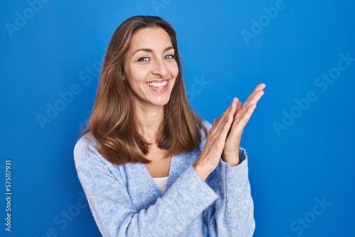 Young woman standing over blue background clapping and applauding happy and joyful, smiling proud hands together