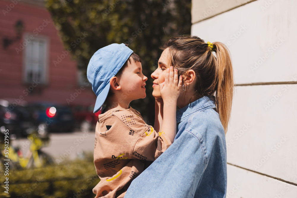 Fototapeta premium Happy mom holding son boy in arms, hugging kid with noses touch, smiling, enjoying warm moment. Younger mum carrying kid and dancing on the street. Motherhood, adoption, family concept.