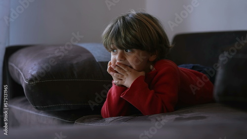 Small child lying on sofa watching cartoons while eating cracker. Candid young boy consuming media online hile relaxing on couch photo