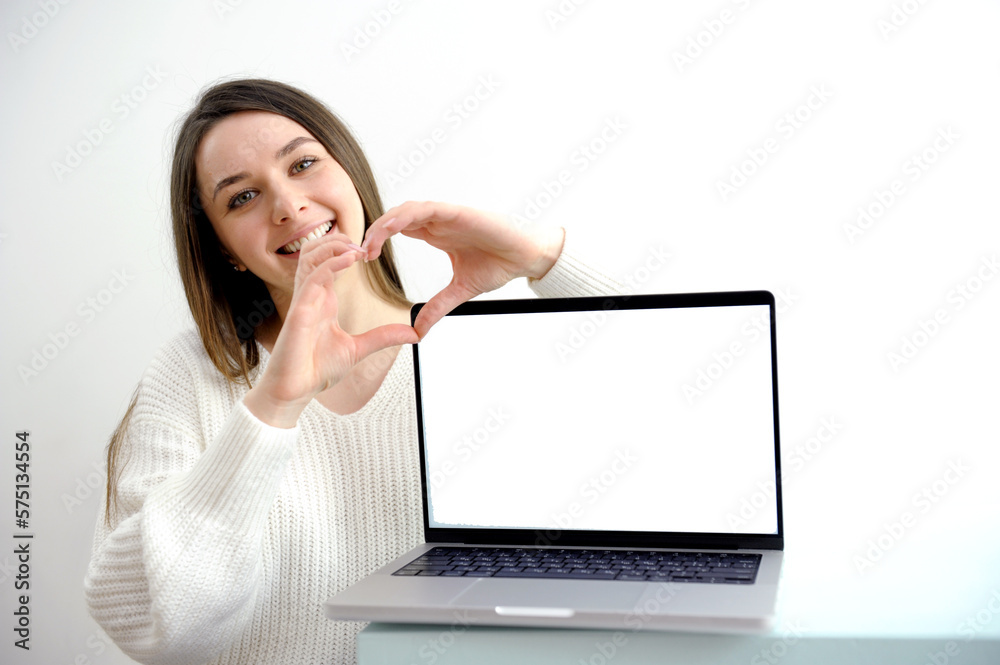 a girl in white shows a heart with her hands smiling near a laptop with an empty space for advertising text of any content girl on a white background points to a laptop screen. High quality photo a girl in white shows a heart with her hands smiling near a laptop with an empty space for advertising text of any content girl on a white background points to a laptop screen. High quality photo