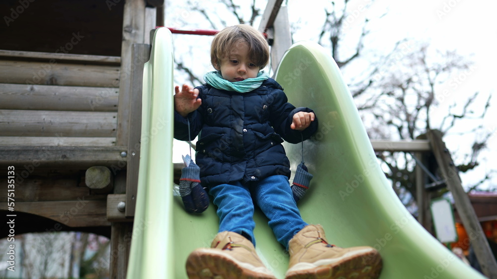 Child sliding down in playground slider during winter season. One little boy wearing coat and scarf outdoors having fun activity Child sliding down in playground slider during winter season. One little boy wearing coat and scarf outdoors having fun activity