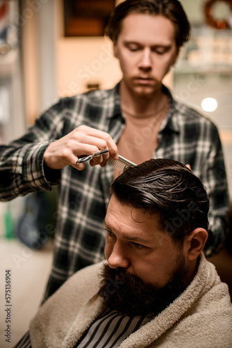 view on handsome bearded man at barbershop and barber cuts and styles his hair