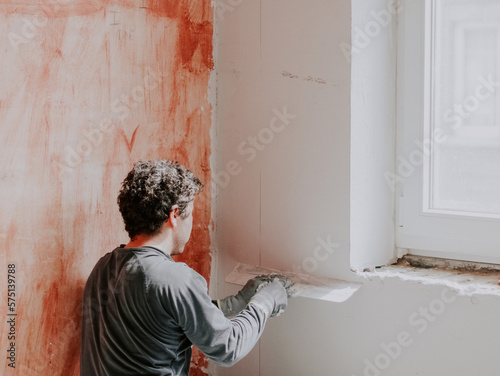 A young caucasian plasterer holds a large trowel and levels a freshly plastered wall © Nataliya
