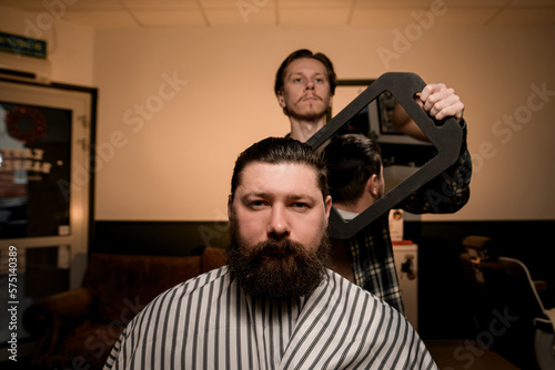portrait of bearded man in a barbershop and barber shows hairstyle with mirror