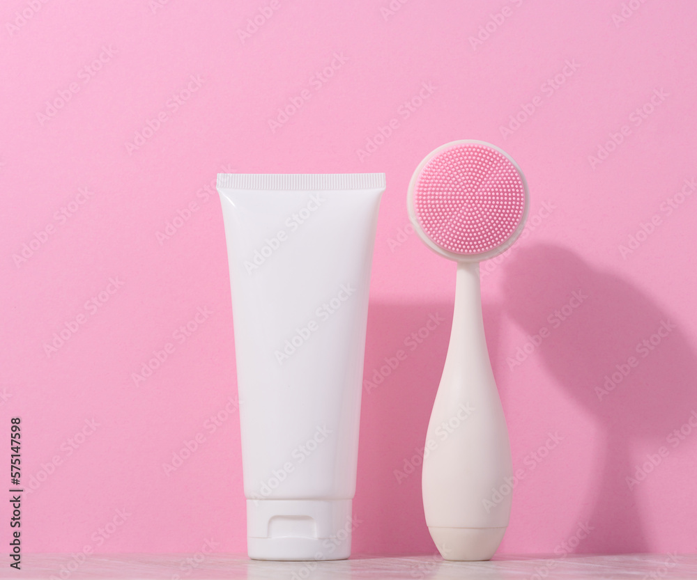 White plastic tube and a massage brush for facial cleansing on a pink background, items for cosmetic procedures White plastic tube and a massage brush for facial cleansing on a pink background, items for cosmetic procedures