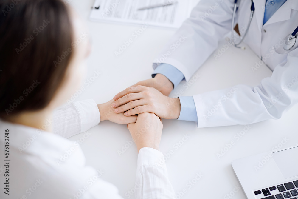 Doctor and patient sitting opposite each other at the table in clinic office. The focus is on female physician's hands reassuring woman, only hands, close up. Medicine concept