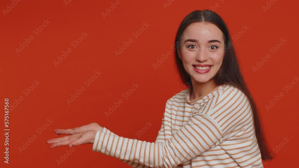Lovely millennial pretty woman showing thumbs up and pointing empty place, advertising area for commercial text, copy space for goods promotion. Young girl. Studio shot indoors on red wall background Lovely millennial pretty woman showing thumbs up and pointing empty place, advertising area for commercial text, copy space for goods promotion. Young girl. Studio shot indoors on red wall background