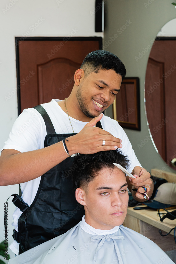 Young Latin American at work providing his services to clients. Barber doing a haircut to a client. Hairdressing service performed by a man. Colombian person working in his business Young Latin American at work providing his services to clients. Barber doing a haircut to a client. Hairdressing service performed by a man. Colombian person working in his business