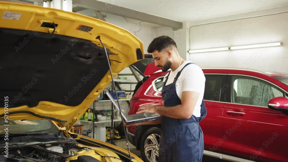 Mechanic man mechanic manager worker using a laptop computer checking car in workshop at auto car repair service center. Engineer young man looking at inspection vehicle details under car hood Mechanic man mechanic manager worker using a laptop computer checking car in workshop at auto car repair service center. Engineer young man looking at inspection vehicle details under car hood