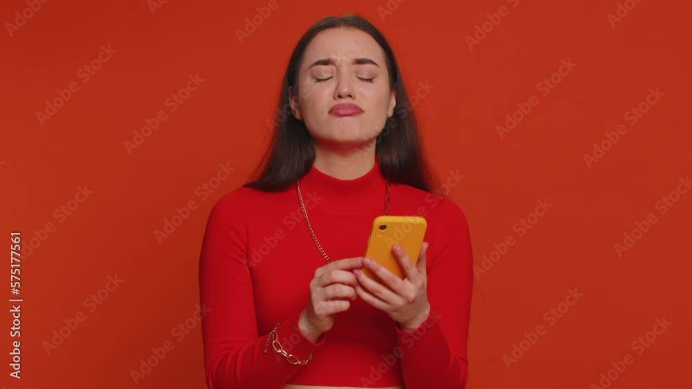Pretty woman playing on smartphone typing browsing, loses becoming surprised sudden lottery results, bad fortune, loss, game fail, bankruptcy. Young girl isolated alone on red studio wall background Pretty woman playing on smartphone typing browsing, loses becoming surprised sudden lottery results, bad fortune, loss, game fail, bankruptcy. Young girl isolated alone on red studio wall background