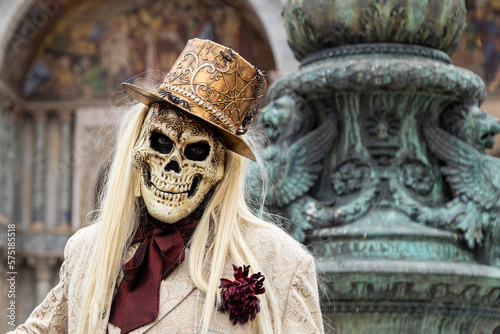 Venice, Veneto, Italy - Feb 19, 2023: Masquerade man in a costume of skull wearing a top hat and a suit during 2023 Venice Carnival celebrations. San Marco Basilica and fountain in the background