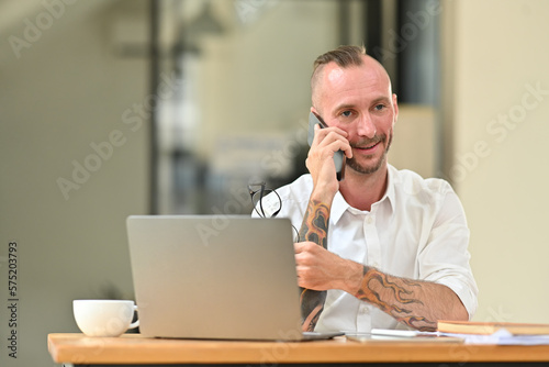 Confident caucasian male manager in a white shirt with tattooed sitting in front of laptop at office desk and talking on mobile phone