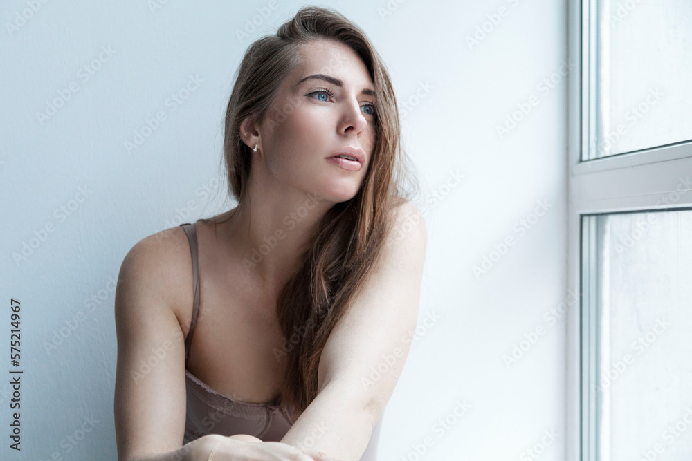 A pensive young woman sits on the windowsill and looks out the window. Loneliness and depression. A pensive young woman sits on the windowsill and looks out the window. Loneliness and depression.