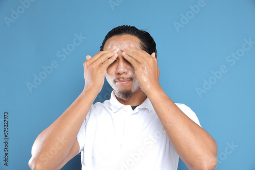 Close up portrait of young Asian man looking at camera, using facial clay mask, posing isolated over blue background. Beauty procedure and skincare routine concept. Horizontol shot photo