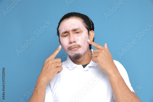 Close up portrait of young Asian man looking at camera, using facial clay mask, posing isolated over blue background. Beauty procedure and skincare routine concept. Horizontol shot photo