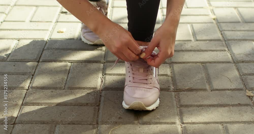 Sportswoman ties laces on shoes for jogging in park illuminated by sunlight. Young woman in black leggings prepares for running in park Sportswoman ties laces on shoes for jogging in park illuminated by sunlight. Young woman in black leggings prepares for running in park
