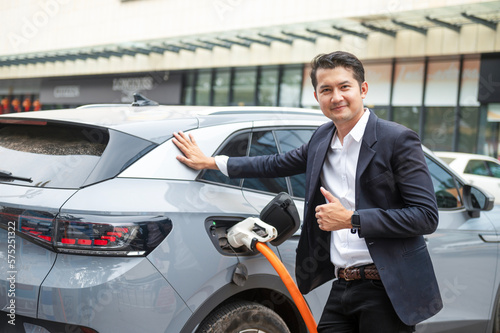 Asian businessman traveling with electric car stopping at charging station standing plugged in internet cable on smartphone smiling joyfully while charging, energy saving electric car view