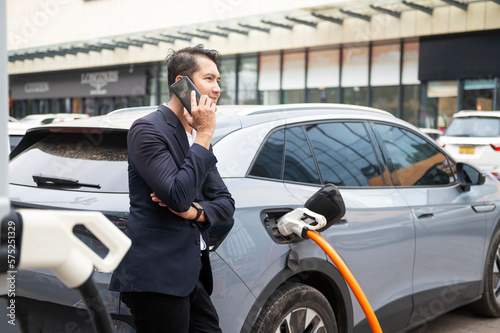 Asian businessman traveling with electric car stopping at charging station standing plugged in internet cable on smartphone smiling joyfully while charging, energy saving electric car view
