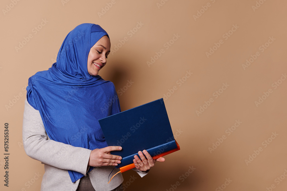 Confident Muslim woman with head covered in hijab, smiling while holding pile of hardcover books in her hands, isolated on beige background with copy space. Portrait of a teacher, professor, educator Confident Muslim woman with head covered in hijab, smiling while holding pile of hardcover books in her hands, isolated on beige background with copy space. Portrait of a teacher, professor, educator