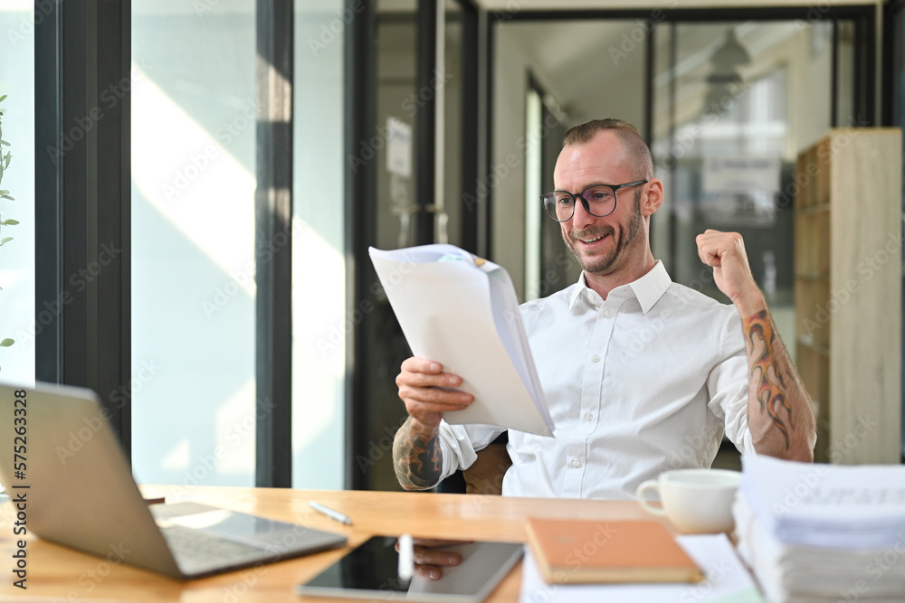 Happy millennial caucasian male employee celebrating success while reading documents in office Happy millennial caucasian male employee celebrating success while reading documents in office