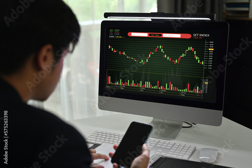 Side view of young male financial analyst using mobile phone and monitoring stocks data candle charts on screen