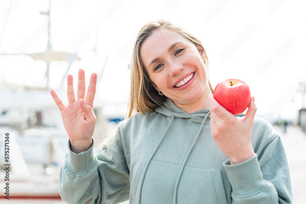 Young blonde woman with an apple at outdoors saluting with hand with happy expression Young blonde woman with an apple at outdoors saluting with hand with happy expression