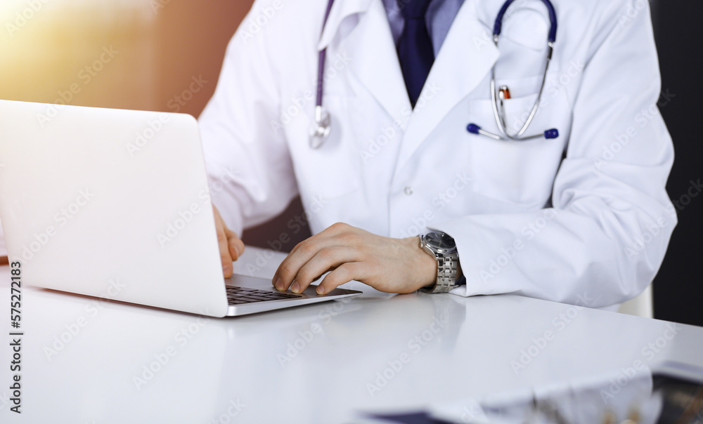 Unknown male doctor sitting and working with laptop in a darkened clinic, glare of light on the background, close-upof hands Unknown male doctor sitting and working with laptop in a darkened clinic, glare of light on the background, close-upof hands