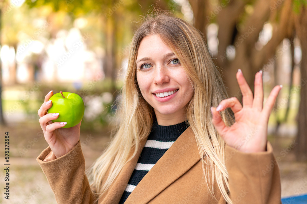 Young pretty blonde woman with an apple at outdoors showing ok sign with fingers Young pretty blonde woman with an apple at outdoors showing ok sign with fingers