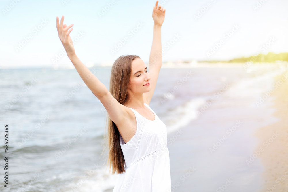 Happy smiling woman in free happiness bliss on ocean beach standing with raising hands. Portrait of a multicultural female model in white summer dress enjoying nature Happy smiling woman in free happiness bliss on ocean beach standing with raising hands. Portrait of a multicultural female model in white summer dress enjoying nature