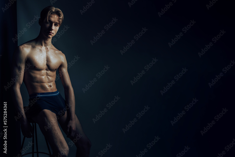 hot male student sit on stool in trunks lean forward and look at camera lens with his topless athletic body with shiny and glossy skin caused by water and sweat with serious, earnest face expression