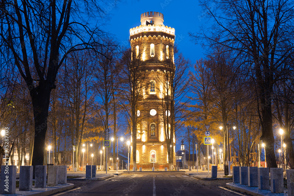 Historic brick water tower built in 1916 in the town of Zaraysk, Moscow region, Russia. View on old water tower against the backdrop of an illuminated evening street with golden lanterns.