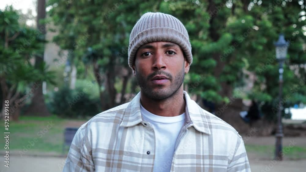 Close up portrait of young attractive african american man with beanie hat looking serious at camera. Front view of a hispanic guy standing in a park outdoors with sad attitude. Slow motion. High Close up portrait of young attractive african american man with beanie hat looking serious at camera. Front view of a hispanic guy standing in a park outdoors with sad attitude. Slow motion. High