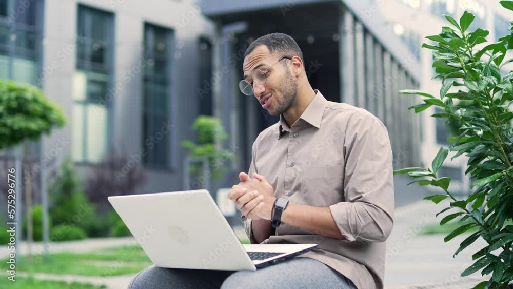 Young adult man talking on a video call using a laptop while sitting on a bench near an office building. Smiling handsome mixed-race male in a shirt and glasses having a conversation with a colleague Young adult man talking on a video call using a laptop while sitting on a bench near an office building. Smiling handsome mixed-race male in a shirt and glasses having a conversation with a colleague
