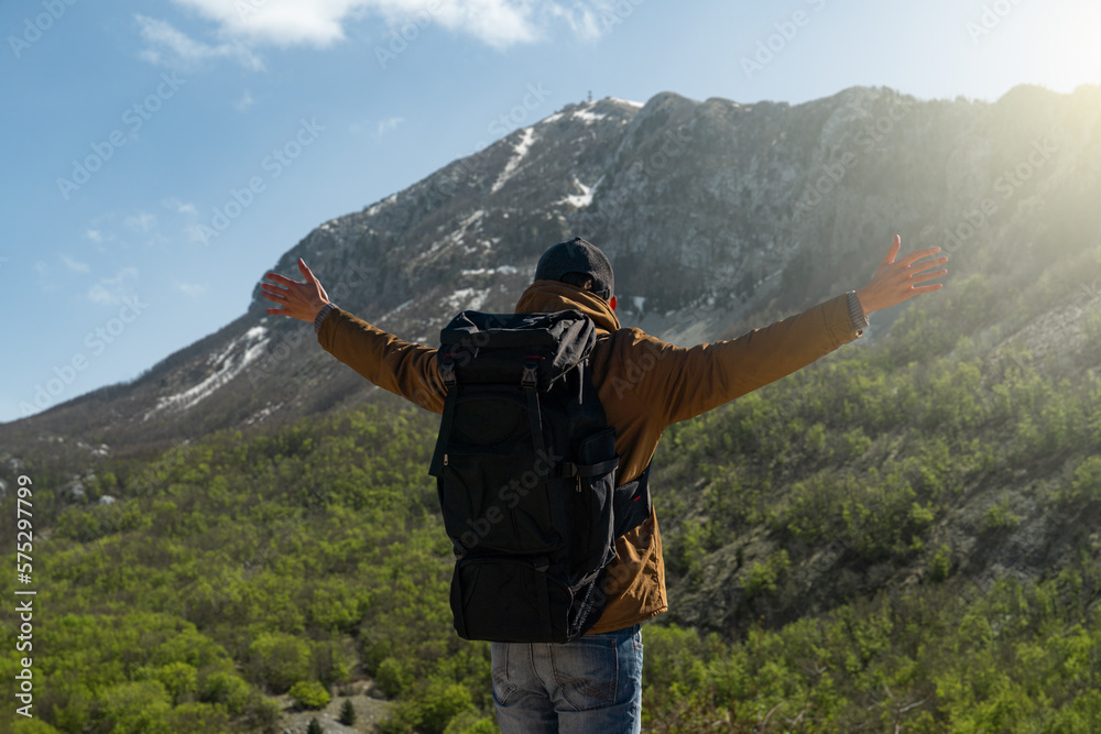 A traveler with a backpack stands on the top of the mountain and holds his hands to the sides	