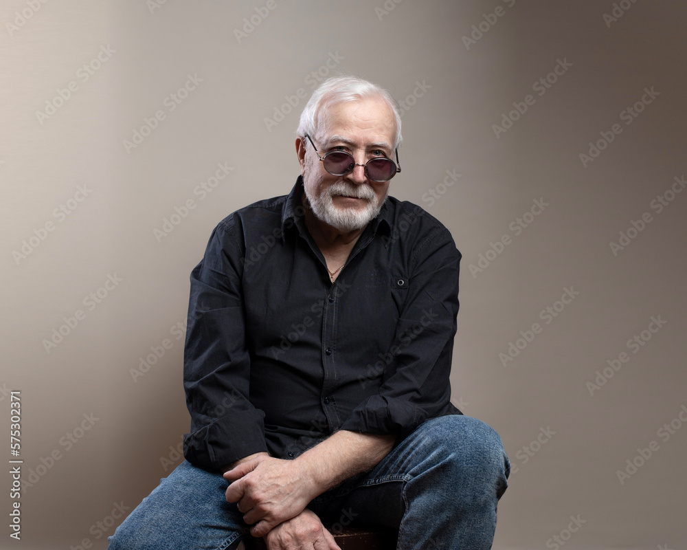 Charismatic elderly gray-haired man in sunglasses and gray shirt posing for camera in studio, sitting on chair on light background Charismatic elderly gray-haired man in sunglasses and gray shirt posing for camera in studio, sitting on chair on light background