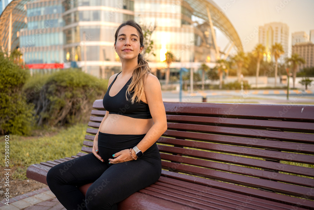 Portrait of friendly young pregnant woman touching her stomach while resting on bench outdoors.