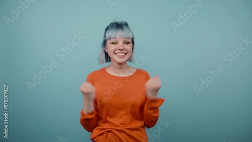 Joyful hipster girl with blue hair stands on a blue background with a happy face raises her hands up and smiles, wears a green sweatshirt